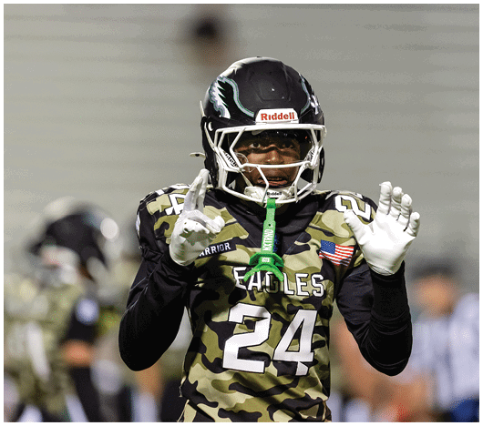 A person wearing a camouflage sports jersey and helmet stands on a football field with arms raised near the goalposts and a scoreboard in the background. Description generated by AI