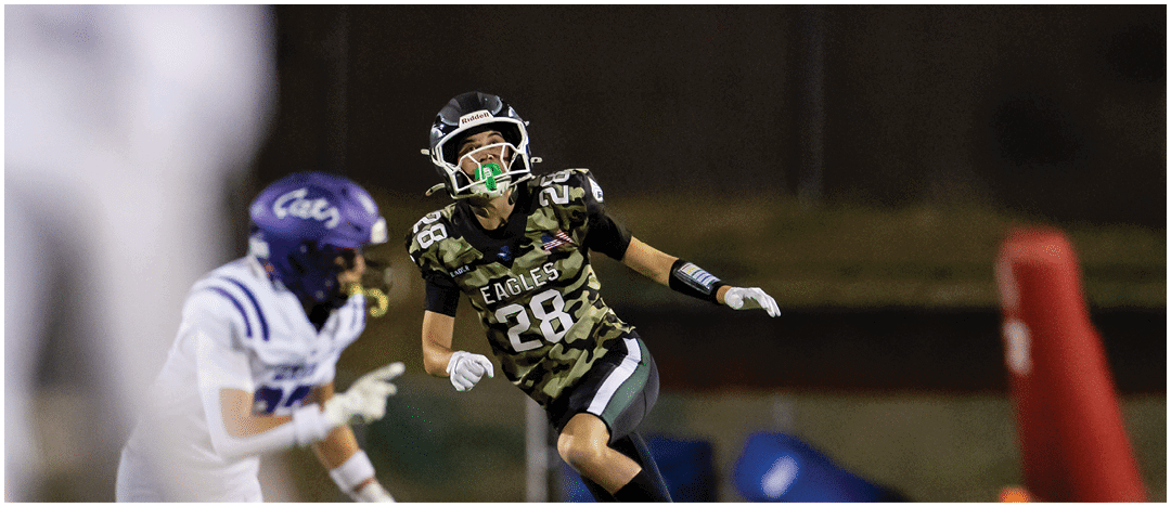 A person wearing a camouflage sports jersey and helmet stands on a football field with arms raised near the goalposts and a scoreboard in the background. Description generated by AI