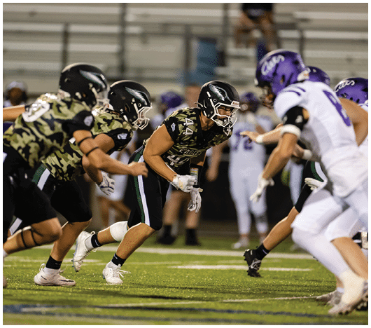 A person wearing a camouflage sports jersey and helmet stands on a football field with arms raised near the goalposts and a scoreboard in the background. Description generated by AI