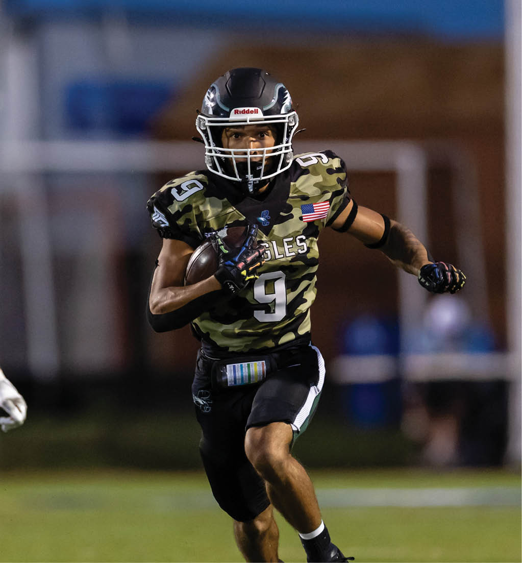 A person wearing a camouflage football uniform with the number nine and an American flag patch is running while holding a football on a field. Description generated by AI