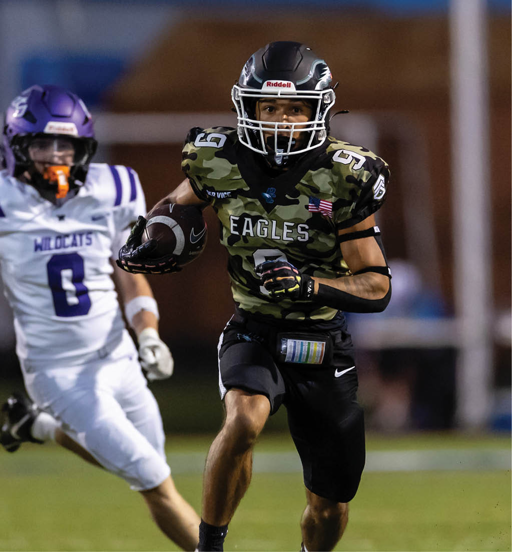A person wearing a camouflage football uniform with the word Eagles is running with a football while another person in a white and purple uniform is reaching out toward them. Description generated by AI