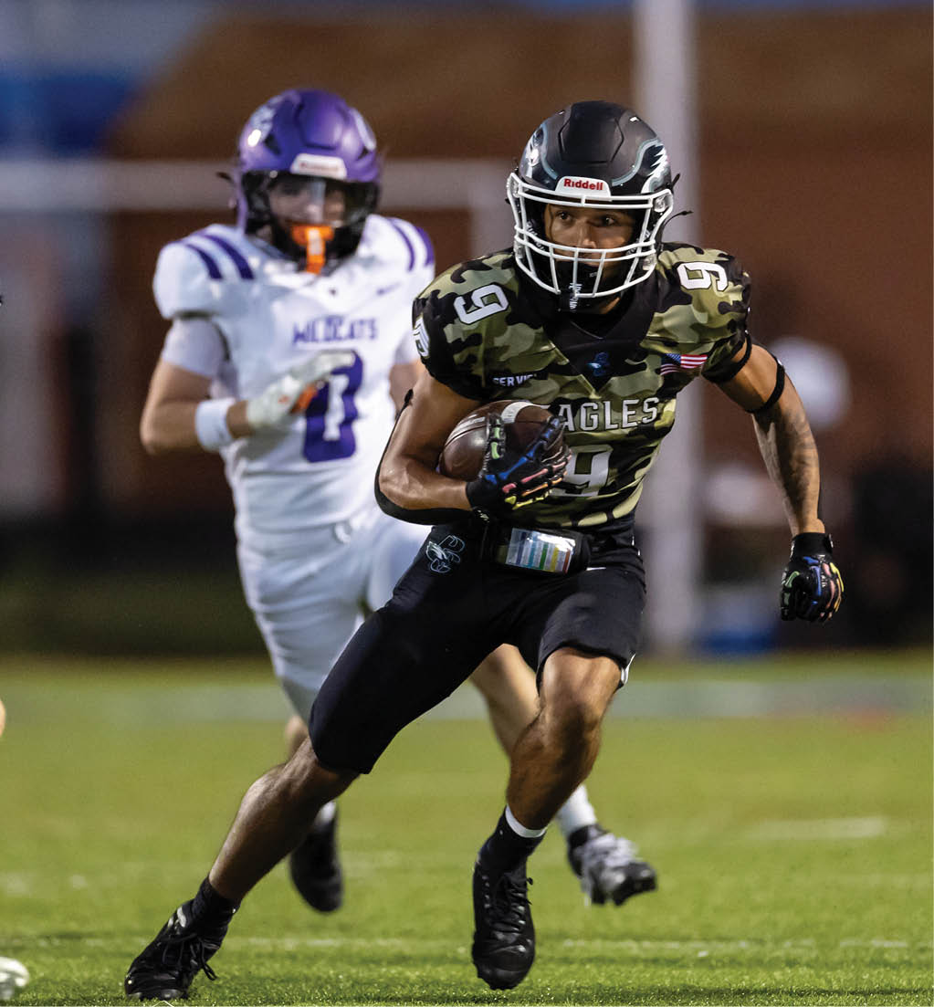 A person wearing a camouflage football uniform with the word Eagles is running with the ball while two people in white and purple football uniforms chase them on a grassy field. Description generated by AI