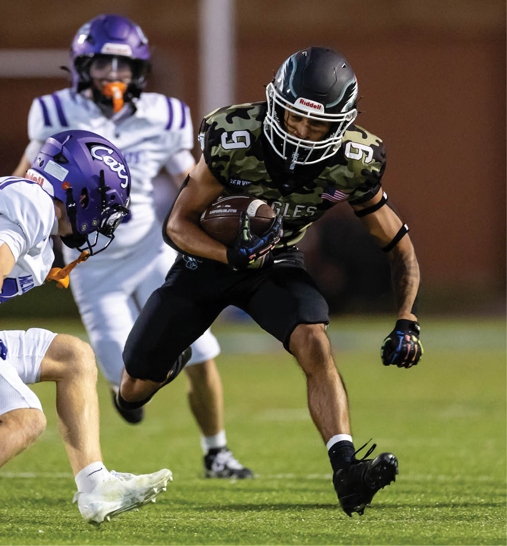 A person in a black and camouflage football uniform is running with the ball while two people in white and purple football uniforms attempt to tackle them on a grassy field. Description generated by AI