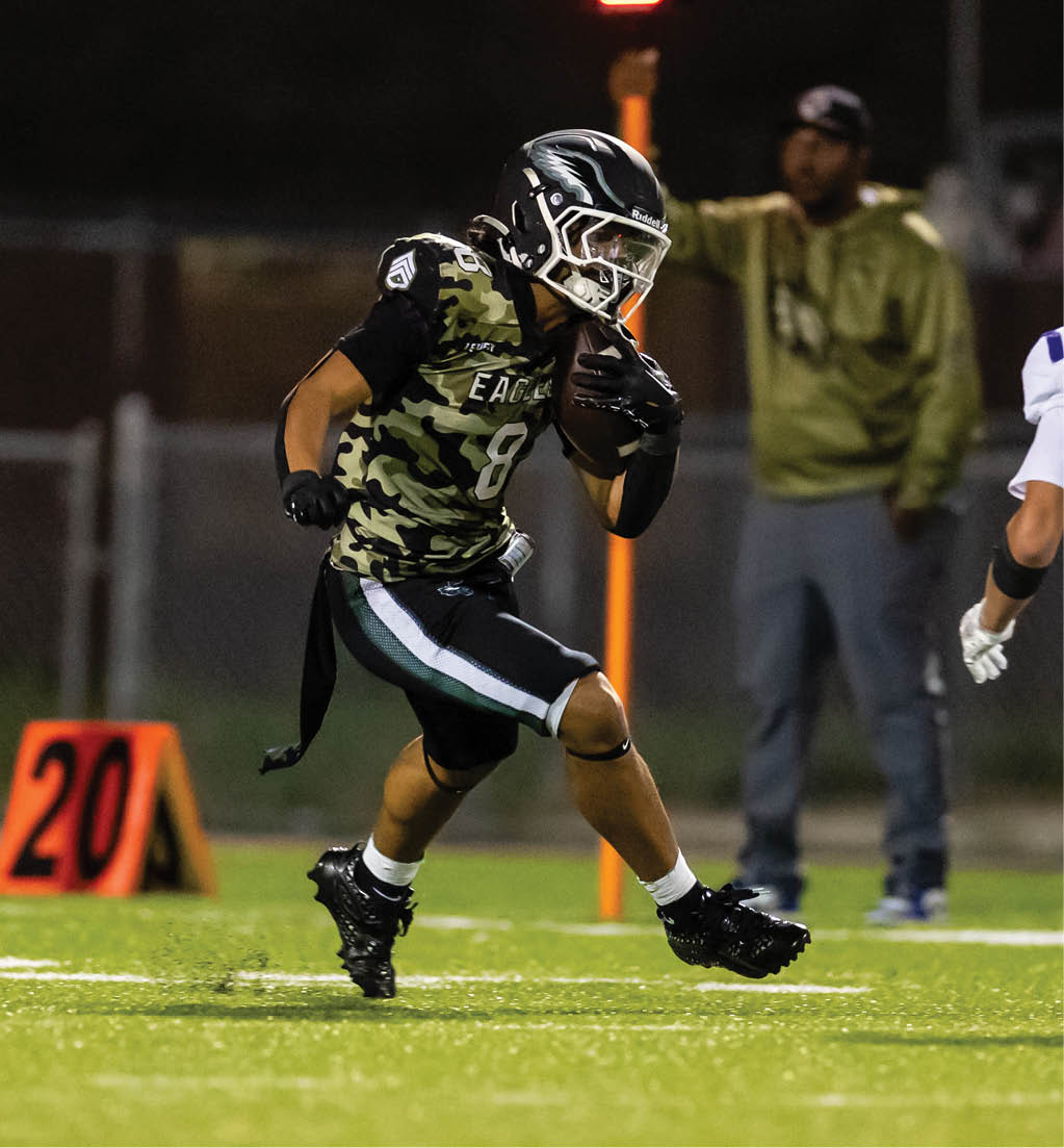 Two people in football uniforms are actively engaged on a field with one holding the ball and the other preparing to defend while a person in casual clothing stands in the background. Description generated by AI