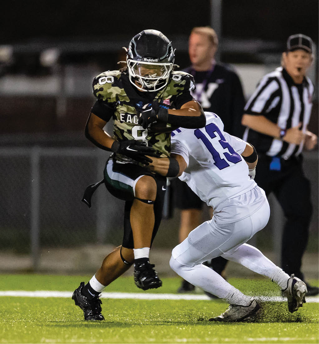 A person in a camouflage football uniform is running with the ball while another person in a white uniform attempts to tackle them on a lit football field with a referee and another person in the background. Description generated by AI