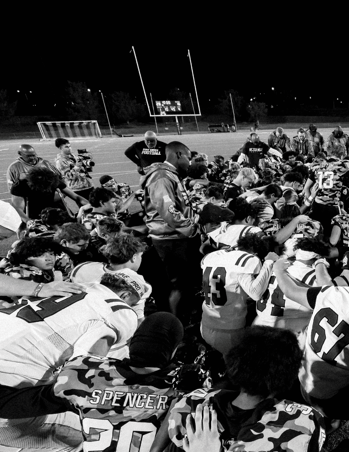 A large group of people in football uniforms and coaches are gathered closely together on a field at night, appearing to be in a team huddle or prayer. Description generated by AI