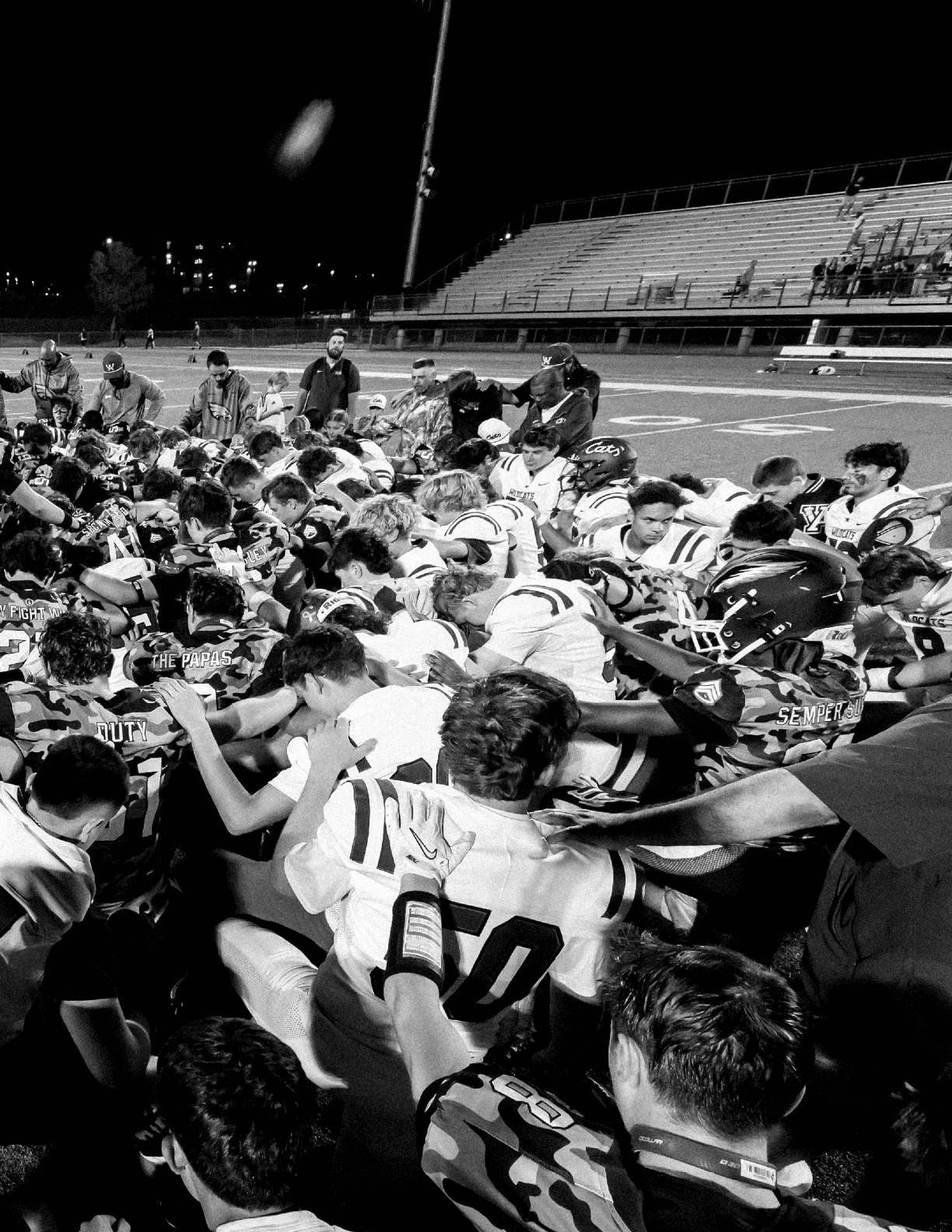 A large group of people in football uniforms are huddled closely together on a field at night, appearing to share a moment of unity or prayer. Description generated by AI
