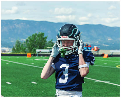 A person wearing a dark blue football uniform with the number three is standing on a green football field adjusting their helmet with mountains and trees in the background. Description generated by AI