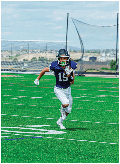 A person wearing a dark sports jersey with the number 19 and white pants is running on a green football field with a mouthguard in their mouth. Description generated by AI