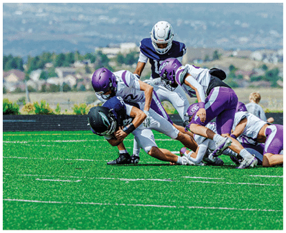 Several people in football uniforms are engaged in a tackle on a green field with a blurred background of houses and hills. Description generated by AI