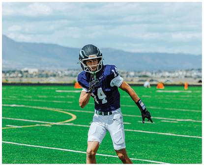 A person wearing a dark blue football uniform and helmet is running on a green football field with mountains and a cloudy sky in the background. Description generated by AI