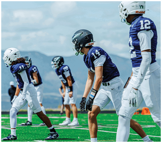 Several people wearing football helmets and uniforms are lined up on a field under a partly cloudy sky. Description generated by AI