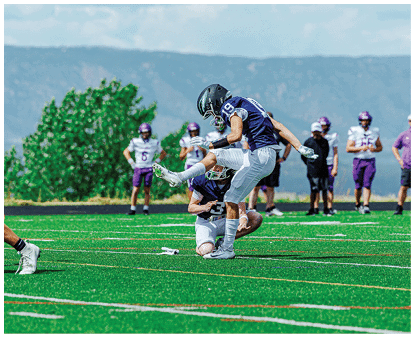 Two people in football uniforms are performing a field goal kick on a green turf field with several people standing in the background near a tree and mountains. Description generated by AI