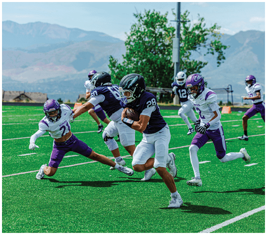 A group of people in football uniforms are actively playing on a green field with mountains and trees in the background. Description generated by AI