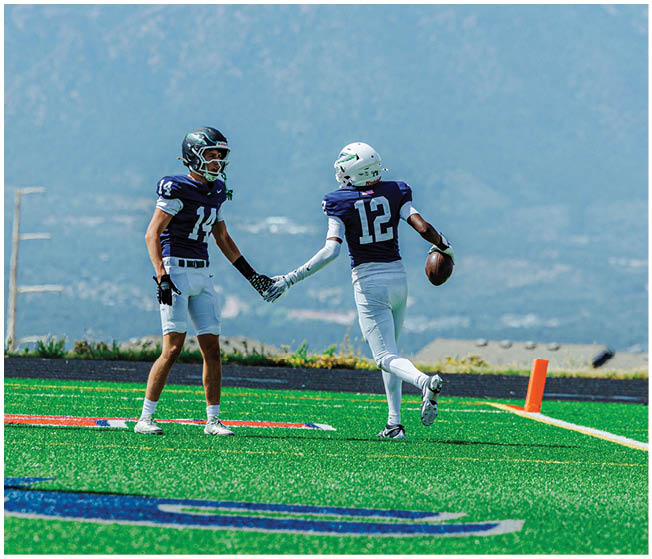 Two people in football uniforms are holding hands on a field near the end zone while other players and a referee stand nearby with mountains in the background. Description generated by AI