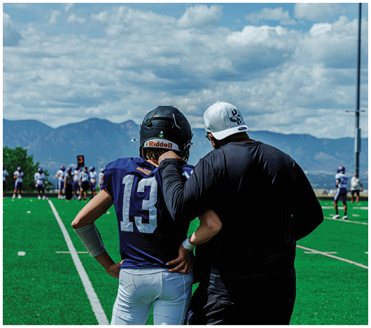 A person in a black long-sleeve shirt and white cap is standing with an arm around a person in a football uniform with number 13 on the back on a green football field with mountains and a cloudy sky in the background. Description generated by AI