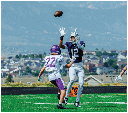 Two people wearing football helmets and gloves are reaching up to catch a football in midair against a backdrop of mountains and a cloudy sky. Description generated by AI