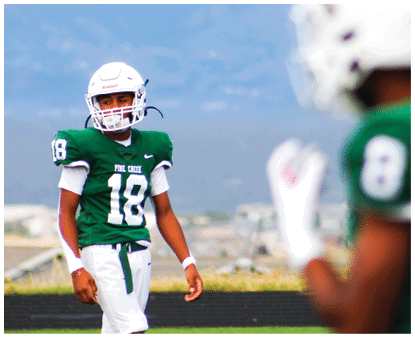 Two people wearing green football uniforms and white helmets are on a field with a blurred background. Description generated by AI