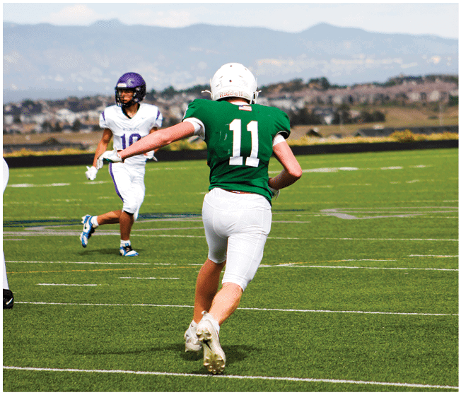Two people wearing football uniforms are on a field with one person in a green jersey running towards the other person in a white jersey against a backdrop of mountains and a partly cloudy sky. Description generated by AI