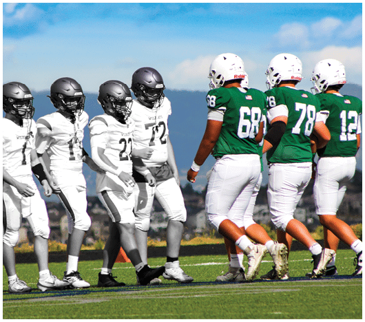 Five people in football uniforms stand on a field facing three other people in green and white football uniforms with helmets under a blue sky. Description generated by AI