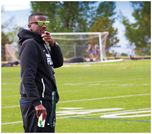 A person wearing a black hoodie and green sunglasses stands on a grassy field near a soccer goal with trees in the background. Description generated by AI
