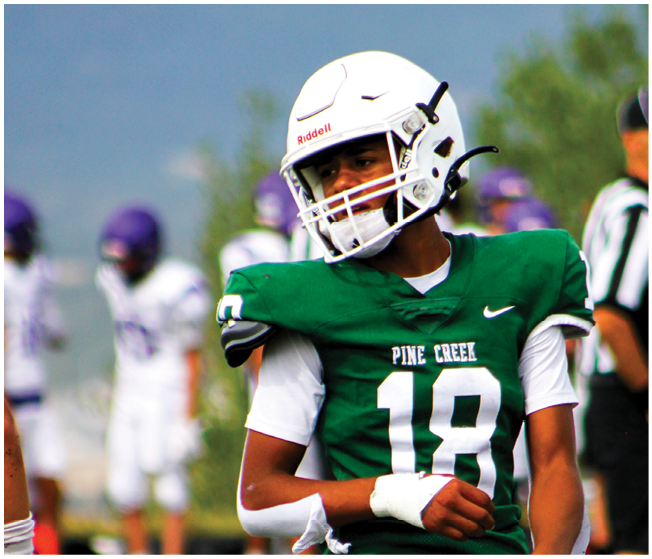 A person wearing a green Pine Creek football jersey and white helmet is standing on a field with blurred players and a referee in the background. Description generated by AI