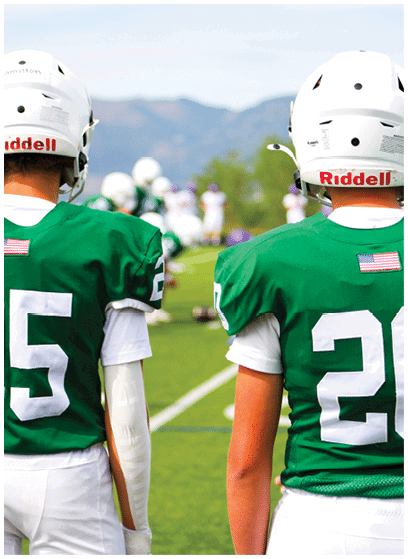 Two people wearing green football jerseys with white numbers and helmets stand on a grassy field facing away from the camera. Description generated by AI