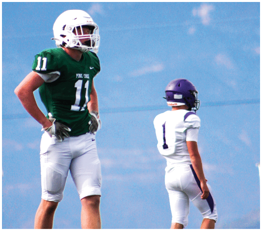 Two people wearing football uniforms and helmets stand on a field with mountains in the background. Description generated by AI