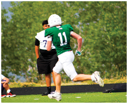 Two people wearing football uniforms are on a field with trees in the background, one in a green jersey with the number 11 running and the other in a white jersey with the number 7 standing behind. Description generated by AI