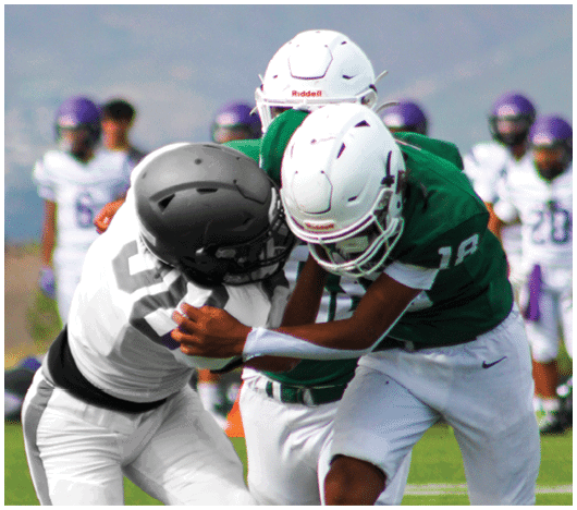 Two people in football uniforms and helmets are engaged in a physical tackle during a game on a grassy field with other players in the background. Description generated by AI