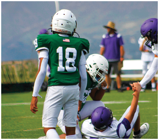 Several people wearing football helmets and uniforms are engaged in a play on a grassy field with one person lying on the ground and others standing or crouching nearby. Description generated by AI