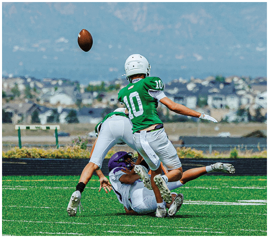 A person wearing a green football jersey with the number 10 and a white helmet is throwing a football outdoors with mountains and buildings in the background. Description generated by AI