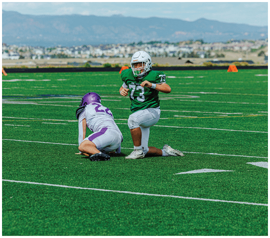 Two children in football uniforms are on a green field with one child holding the ball and the other kneeling in front of them. Description generated by AI