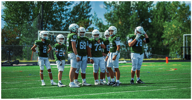 Seven people wearing green football jerseys and helmets stand together on a grassy field with trees and mountains in the background. Description generated by AI