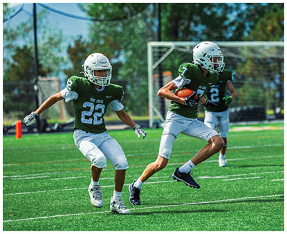 Three people in green and white football uniforms are running on a grassy field with one person holding a football. Description generated by AI