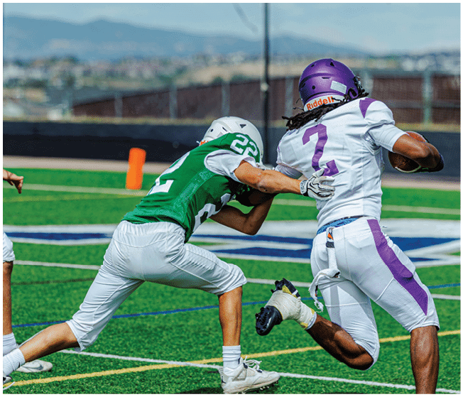 Three people in football uniforms are on a field with one person in a white and purple uniform running with the ball while two others in green and white uniforms attempt to tackle. Description generated by AI