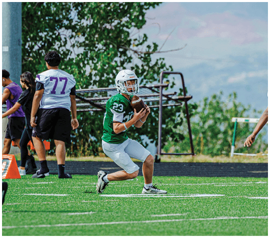 A person in a green jersey is holding a football and preparing to throw it while another person in a white and purple jersey approaches on a grassy field with other people and equipment in the background. Description generated by AI