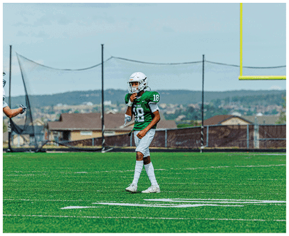 A person in a green football uniform with the number 18 is standing on a field near a yellow goalpost with a cityscape in the background. Description generated by AI