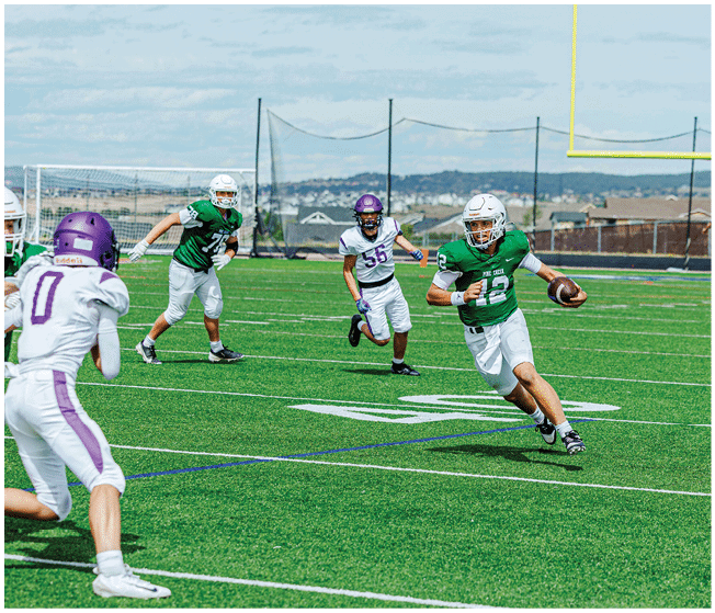 A person in a green jersey is running with a football while three people in white and purple jerseys are chasing on a green football field with a goalpost and net in the background. Description generated by AI