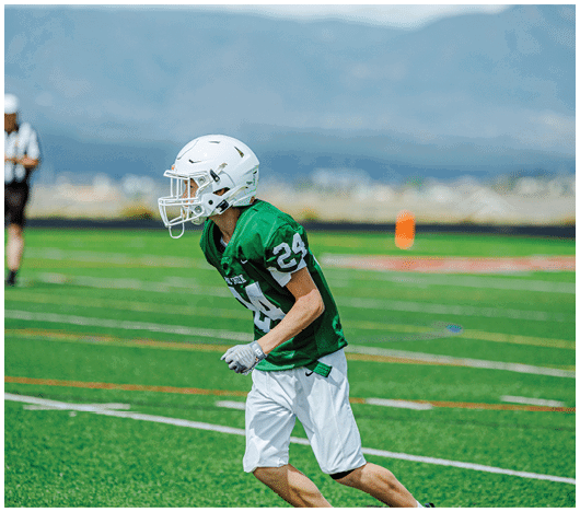 A person wearing a green football jersey and white helmet is running on a grassy football field with mountains in the background. Description generated by AI