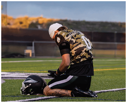 A person wearing a camouflage sports jersey and kneeling on a football field with a helmet placed on the ground in front of them. Description generated by AI