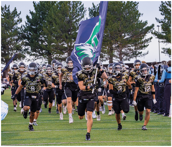 A group of football players in black and camouflage uniforms are running on a field with one player holding a large flag featuring an eagle logo. Description generated by AI