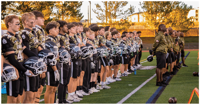 A line of people in football uniforms and helmets stand side by side on a field facing a line of people in matching jackets holding helmets during sunset. Description generated by AI
