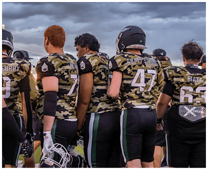 People wearing camouflage football uniforms stand together on a field under a cloudy sky, some holding helmets. Description generated by AI