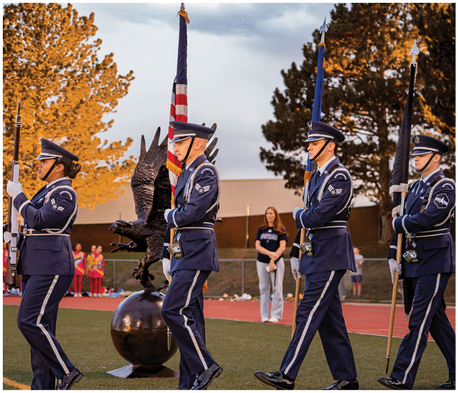 Three people in formal military uniforms march on a field carrying flags near a large eagle statue while children and adults stand in the background. Description generated by AI