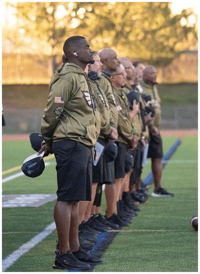 A group of people in sports uniforms stand in a line on a field while one person in casual clothing appears to be moving or gesturing near them. Description generated by AI
