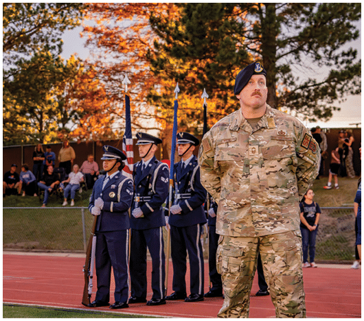 A person in military uniform stands in the foreground while three people in formal dress uniforms hold flags on a track field with spectators and autumn trees in the background. Description generated by AI