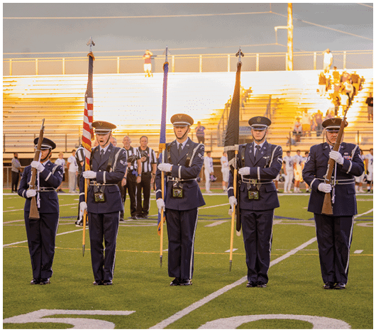Four people in formal military uniforms stand on a football field holding rifles and flags with referees and other people in the background. Description generated by AI