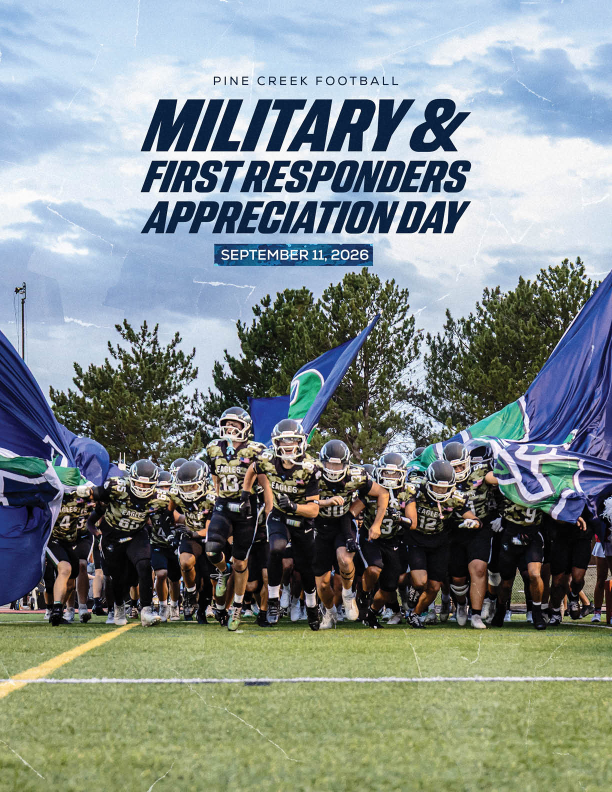 A group of football players in black uniforms run onto a field holding large blue and green flags under a cloudy sky with text above announcing Military and First Responders Appreciation Day on September 11, 2026. Description generated by AI