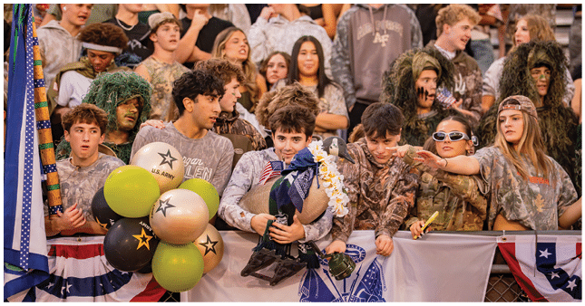 A group of people wearing camouflage clothing and holding balloons and a bouquet are gathered behind a decorated barrier at an outdoor event. Description generated by AI